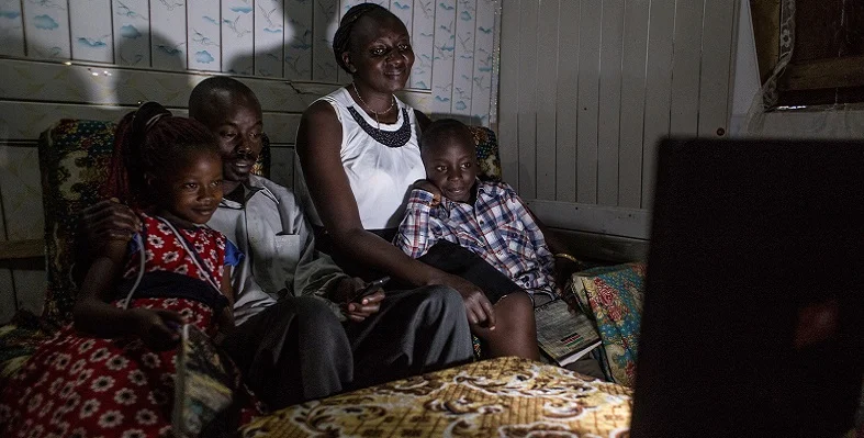 A Kenyan family sat on the sofa watching and illuminated by a TV